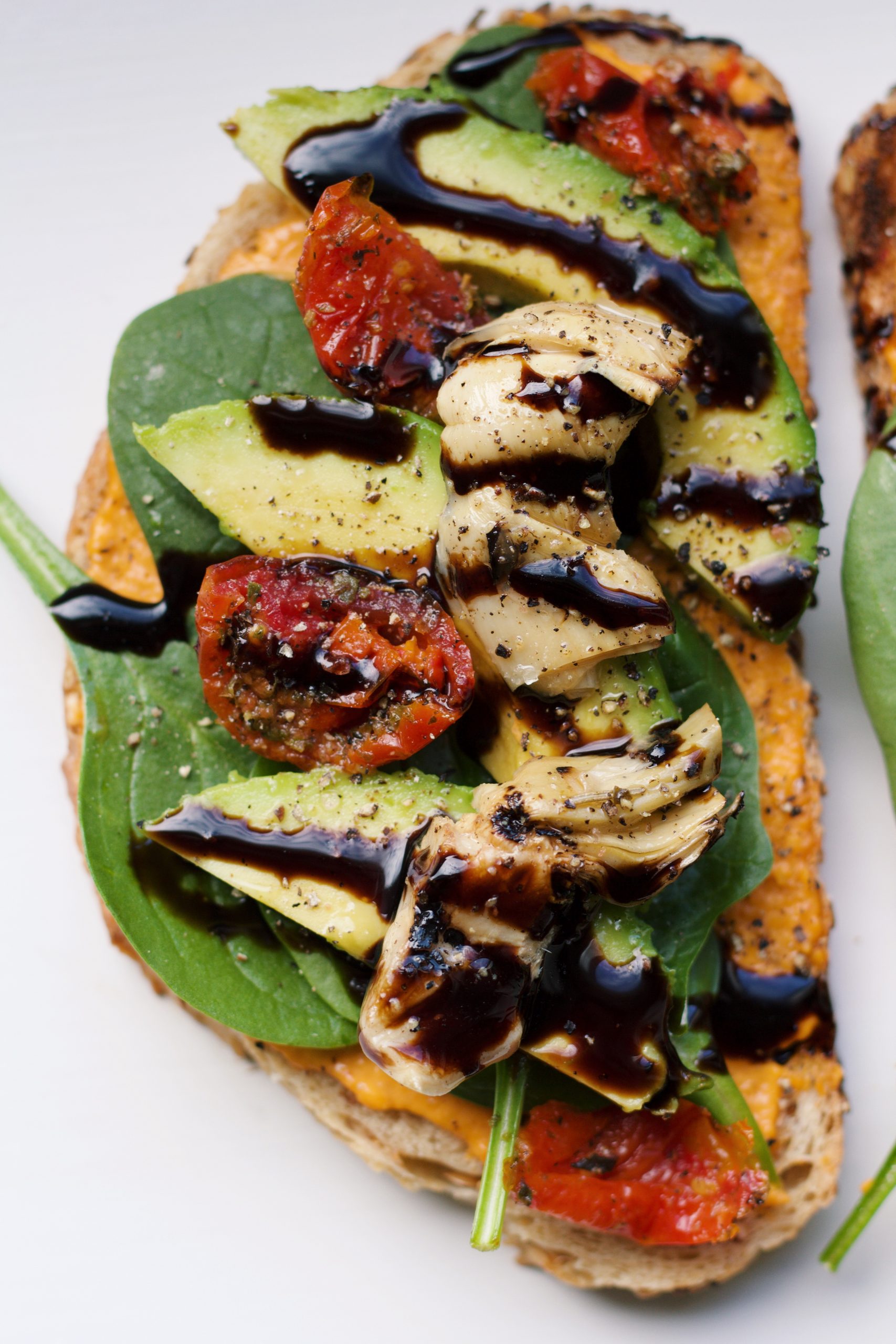 Toast met hummus, baby spinazie, avocado, semi-zongedroogde tomaten, artisjok en balsamico crème